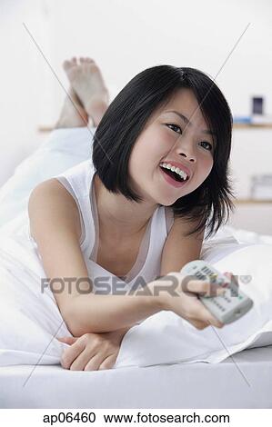 Stock Image - Young woman with TV remote control, smiling. Fotosearch
