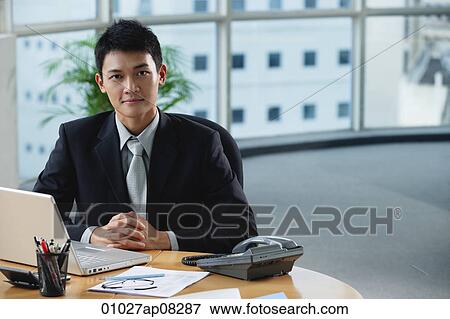 Stock Photo - A man looks at the camera as he sits at his desk. Fotosearch
