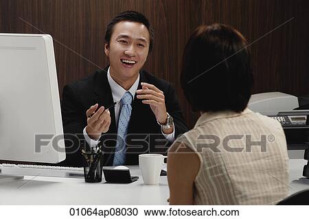 Stock Image - A man sits at his desk while he talks with a woman at work. Fotosearch