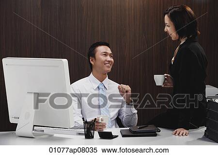 Stock Photography - A man sits at his desk while he talks with a woman at work. Fotosearch