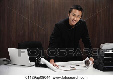 Picture - A man smiles at the camera as he rolls out plans on his desk. Fotosearch