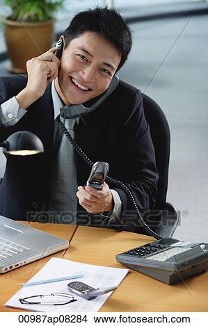 A man smiles at the camera as he sits at his desk which has lots of phones on it View Large Photo Image Picture - A man smiles at the camera as he sits at his desk which has lots of phones on it. Fotosearch