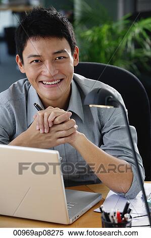 Stock Photography - A man smiles at the camera as he sits at his desk. Fotosearch
