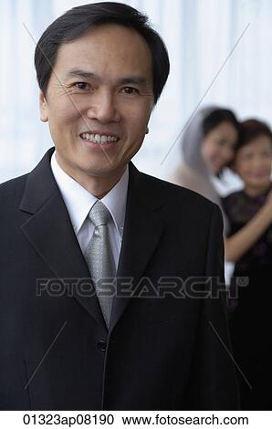 A man smiles at the camera while a bride and her mother hug in the background View Large Photo Image Stock Image - A man smiles at the camera while a bride and her mother hug in the background. Fotosearch