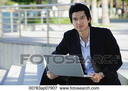 Stock Photo - A man uses his laptop outdoors while he looks at the camera. Fotosearch