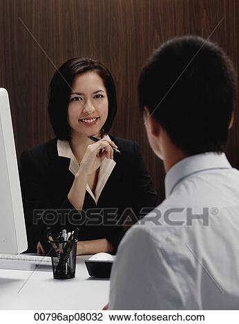 A woman sits at her desk while she talks with a man View Large Photo Image Stock Image - A woman sits at her desk while she talks with a man. Fotosearch