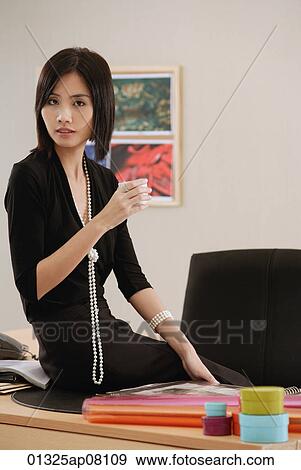 A woman sits on her desk while she looks at the camera View Large Photo Image Stock Photo - A woman sits on her desk while she looks at the camera. Fotosearch