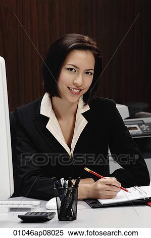 Stock Photography - A woman smiles at the camera while she works at her desk. Fotosearch