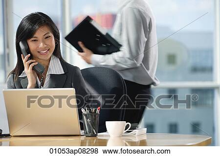 Stock Photo - A woman talks on the phone as she sits at her desk. Fotosearch