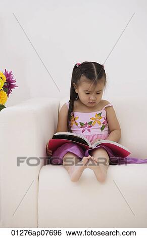 Stock Photograph - A young girl sitting on the couch with a book. Fotosearch