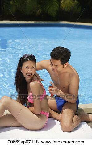 Couple by sitting by swimming pool, man applying sun tan lotion on woman's back View Large Photo Image Stock Photo - Couple by sitting by swimming pool, man applying sun tan lotion on woman's back. Fotosearch