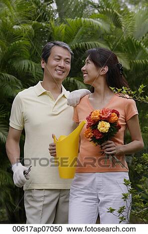 Stock Image - Couple in garden, woman holding bouquet of flowers and watering can, smiling at man next to her. Fotosearch