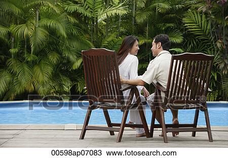 Stock Image - Couple sitting by swimming pool, looking at each other, rear view. Fotosearch