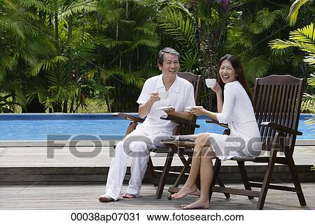 Couple sitting outdoors by swimming pool, having drinks View Large Photo Image Stock Image - Couple sitting outdoors by swimming pool, having drinks. Fotosearch