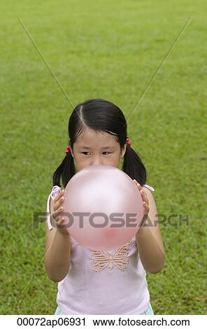 Stock Image - Girl standing on grass, blowing up pink balloon. Fotosearch