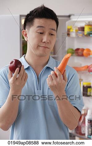 Man holding an apple and a carrot, looking frustrated View Large Photo Image Stock Photo - Man holding an apple and a carrot, looking frustrated. Fotosearch
