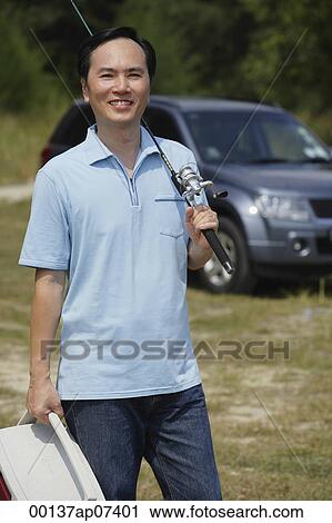 Stock Image - Man holding fishing pole over shoulder and cooler, car in background. Fotosearch