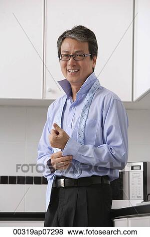 Man in kitchen, buttoning his shirt and tie loose around neck. smiling at camera. Getting ready for work. View Large Photo Image Stock Photo - Man in kitchen, buttoning his shirt and tie loose around neck. smiling at camera. Getting ready for work.. Fotosearch