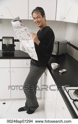 Man in kitchen, leaning on counter, holding newspaper and looking at camera smiling. View Large Photo Image Picture - Man in kitchen, leaning on counter, holding newspaper and looking at camera smiling.. Fotosearch