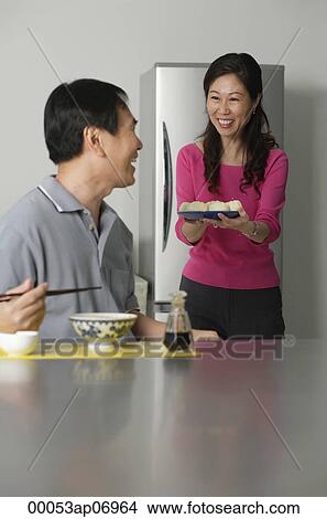 Mature couple in kitchen, man sitting in front of a bowl of noodles, woman behind him holding a plate of food View Large Photo Image Picture - Mature couple in kitchen, man sitting in front of a bowl of noodles, woman behind him holding a plate of food. Fotosearch