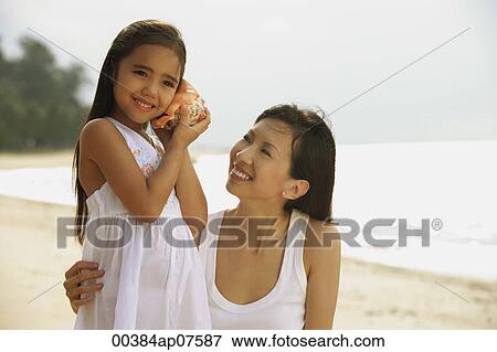 Stock Photo - Mother and daughter on beach, daughter with conk shell to ear listening to ocean. Fotosearch