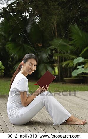 Stock Image - side profile of woman sitting on porch in tropical setting, writing in journal, diary, and looking at camera, smiling. Fotosearch