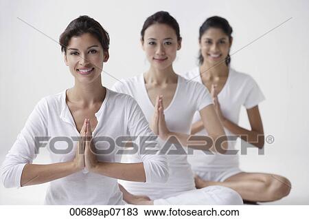 Stock Image - three women of mixed race sitting with legs crossed, arms in namaste, smiling at camera. Fotosearch