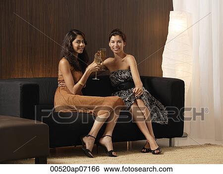 Two girls sitting on couch holding champagne, toasting, smiling at camera View Large Photo Image Stock Photograph - Two girls sitting on couch holding champagne, toasting, smiling at camera. Fotosearch