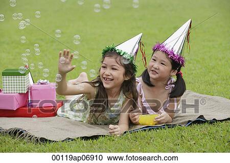 Stock Image - Two girls wearing party hats lying on picnic blanket, looking at bubbles. Fotosearch