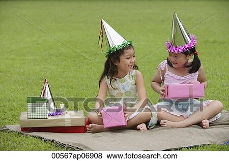 Stock Photo - Two girls wearing party hats sitting on picnic blanket, holding pink gift boxes. Fotosearch