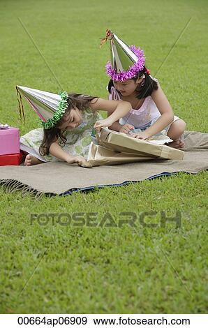 Two girls wearing party hats sitting on picnic blanket, opening a gift View Large Photo Image Stock Photo - Two girls wearing party hats sitting on picnic blanket, opening a gift. Fotosearch