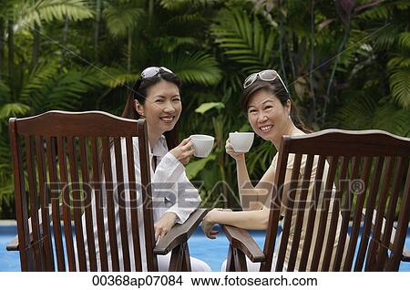 Picture - Two women sitting side by side outdoors, holding cups, turning to look at camera. Fotosearch