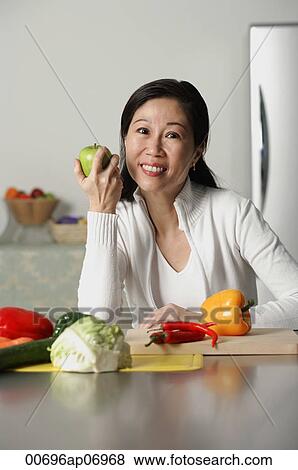 Stock Photo - Woman in kitchen, holding an apple, vegetables on table around her. Fotosearch