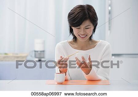 Stock Photo - Woman in kitchen, holding bowl and chopsticks, eating. Fotosearch
