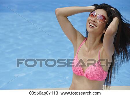 Stock Image - Woman in pink bikini, sitting by swimming pool, hands in hair, smiling. Fotosearch