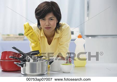 Stock Photo - Woman leaning on kitchen counter, holding cleaning sponge, stack of pots and pans in front of her. Fotosearch