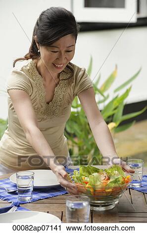 Stock Image - Woman setting a table for a meal, putting bowl of salad on the table. Fotosearch