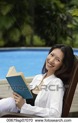 Woman sitting by swimming pool, holding a book, smiling at camera View Large Photo Image Stock Image - Woman sitting by swimming pool, holding a book, smiling at camera. Fotosearch