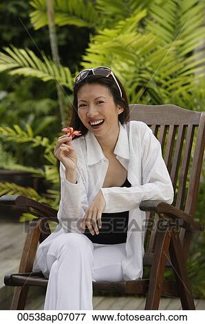 Stock Photo - Woman sitting in garden, holding flower, smiling. Fotosearch