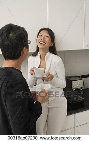 Woman sitting on counter in kitchen laughing with man facing her and his back towards camera. both holding coffee cups. View Large Photo Image Stock Image - Woman sitting on counter in kitchen laughing with man facing her and his back towards camera. both holding coffee cups.. Fotosearch
