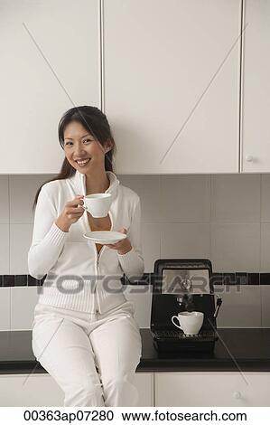 Stock Image - Woman sitting on counter in kitchen next to cappuccino machine, holding coffee, smiling, looking at camera.. Fotosearch