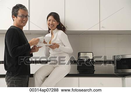 Woman sitting on counter in kitchen with man standing next to her, holding coffee cups, smiling and looking at camera. View Large Photo Image Stock Photo - Woman sitting on counter in kitchen with man standing next to her, holding coffee cups, smiling and looking at camera.. Fotosearch