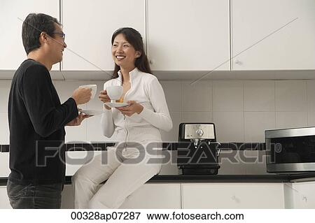 Woman sitting on counter in kitchen with man standing next to her, looking at each other, holding coffee cups and smiling View Large Photo Image Stock Photo - Woman sitting on counter in kitchen with man standing next to her, looking at each other, holding coffee cups and smiling. Fotosearch