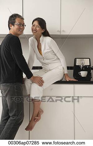 Woman sitting on counter in kitchen with man leaning into her, looking at camera, smiling View Large Photo Image Stock Photography - Woman sitting on counter in kitchen with man leaning into her, looking at camera, smiling. Fotosearch