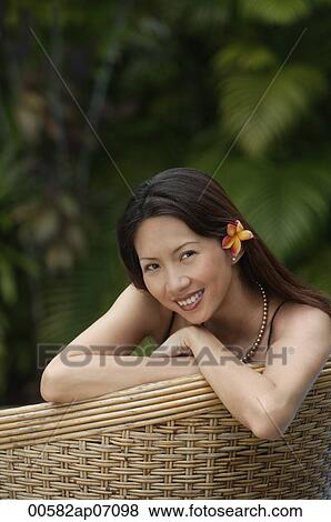 Stock Photo - Woman sitting on rattan chair, wearing flower, portrait. Fotosearch