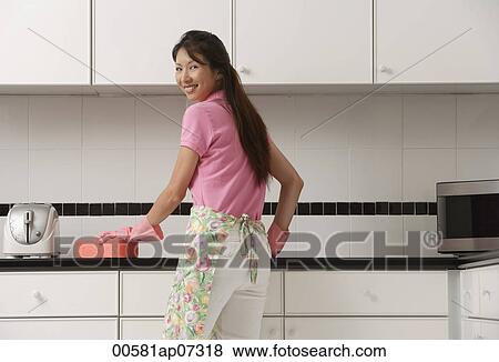 woman standing in kitchen, wearing gloves and apron and cleaning counter with big pink sponge, looking over shoulder at camera, smiling. View Large Photo Image Stock Photo - woman standing in kitchen, wearing gloves and apron and cleaning counter with big pink sponge, looking over shoulder at camera, smiling.. Fotosearch