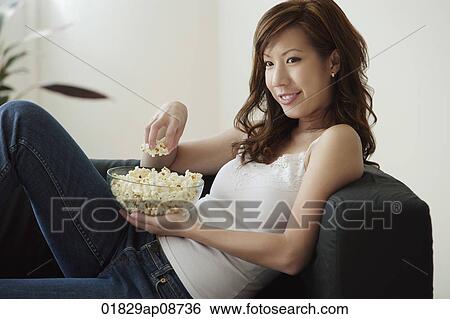 Stock Photograph - Young woman eating popcorn on sofa. Fotosearch