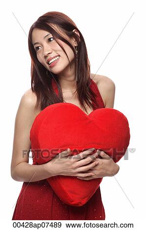 Stock Photo - Young woman holding red heart shaped pillow, looking away. Fotosearch