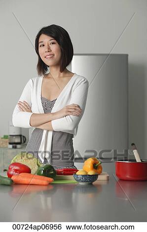 Stock Photograph - Young woman in kitchen, arms crossed, vegetables on the table in front of her. Fotosearch