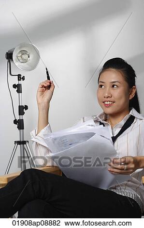 Stock Image - Young woman sitting with paper and pen looking into distance. Fotosearch